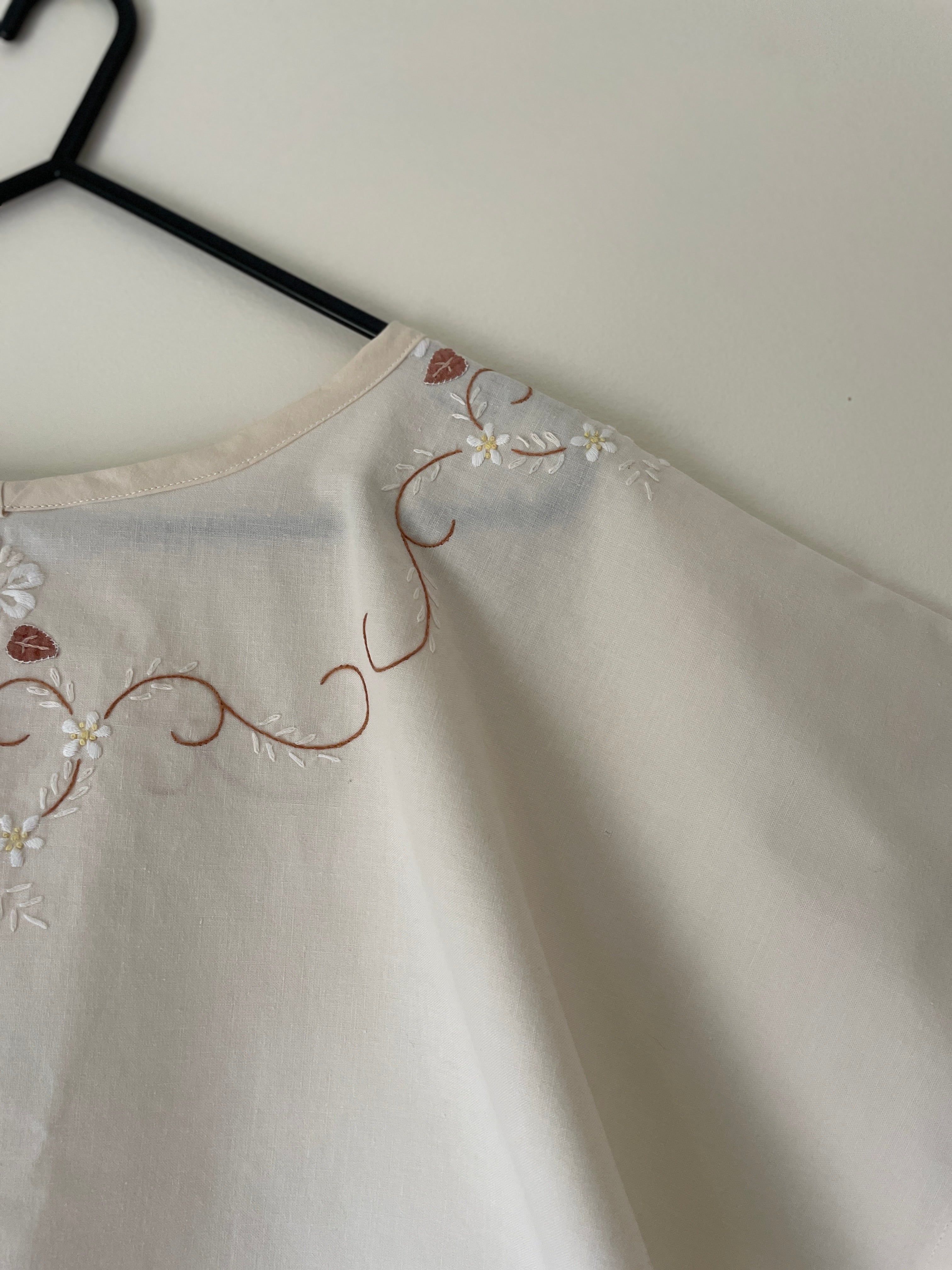 Light-colored top with floral embroidery on a hanger against a white background