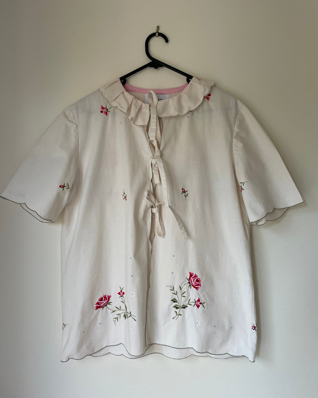 White blouse with floral embroidery on a hanger against a plain wall