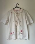White blouse with floral embroidery on a hanger against a plain wall