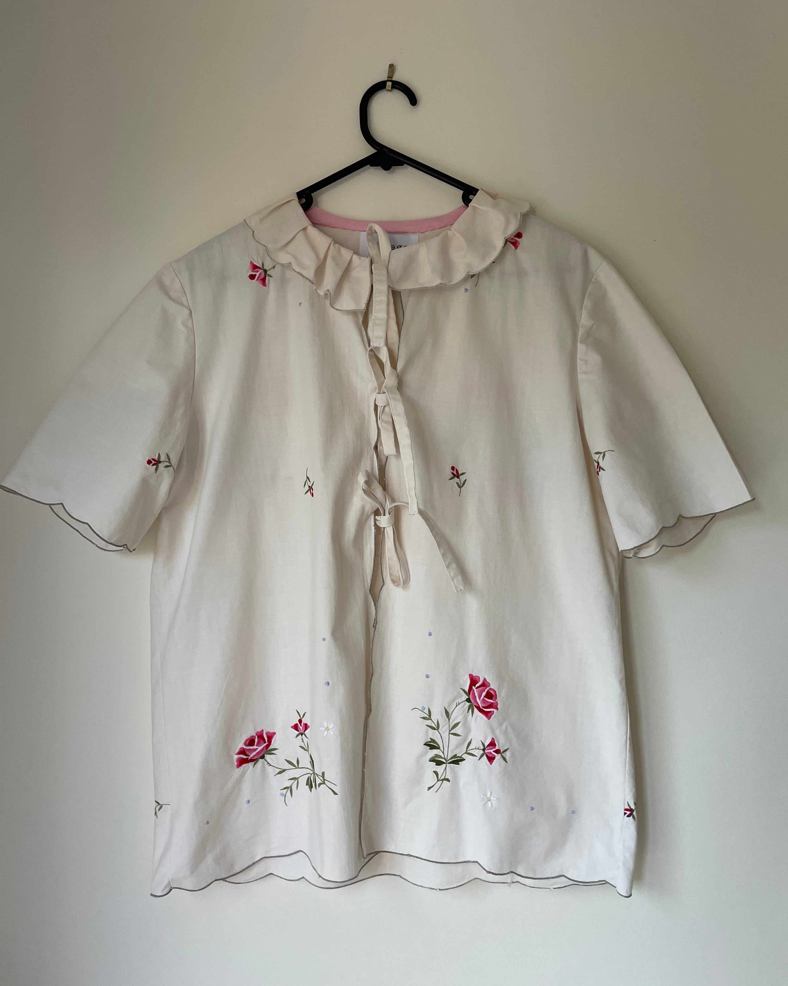 White blouse with floral embroidery on a hanger against a plain wall