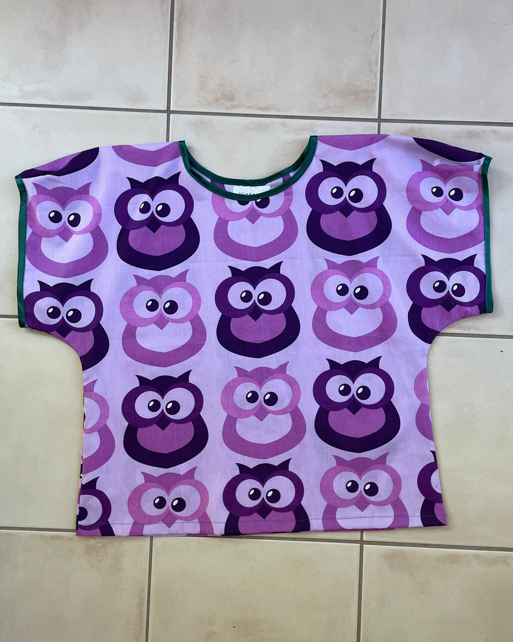 Purple t-shirt with owl pattern on a wooden hanger against a beige background