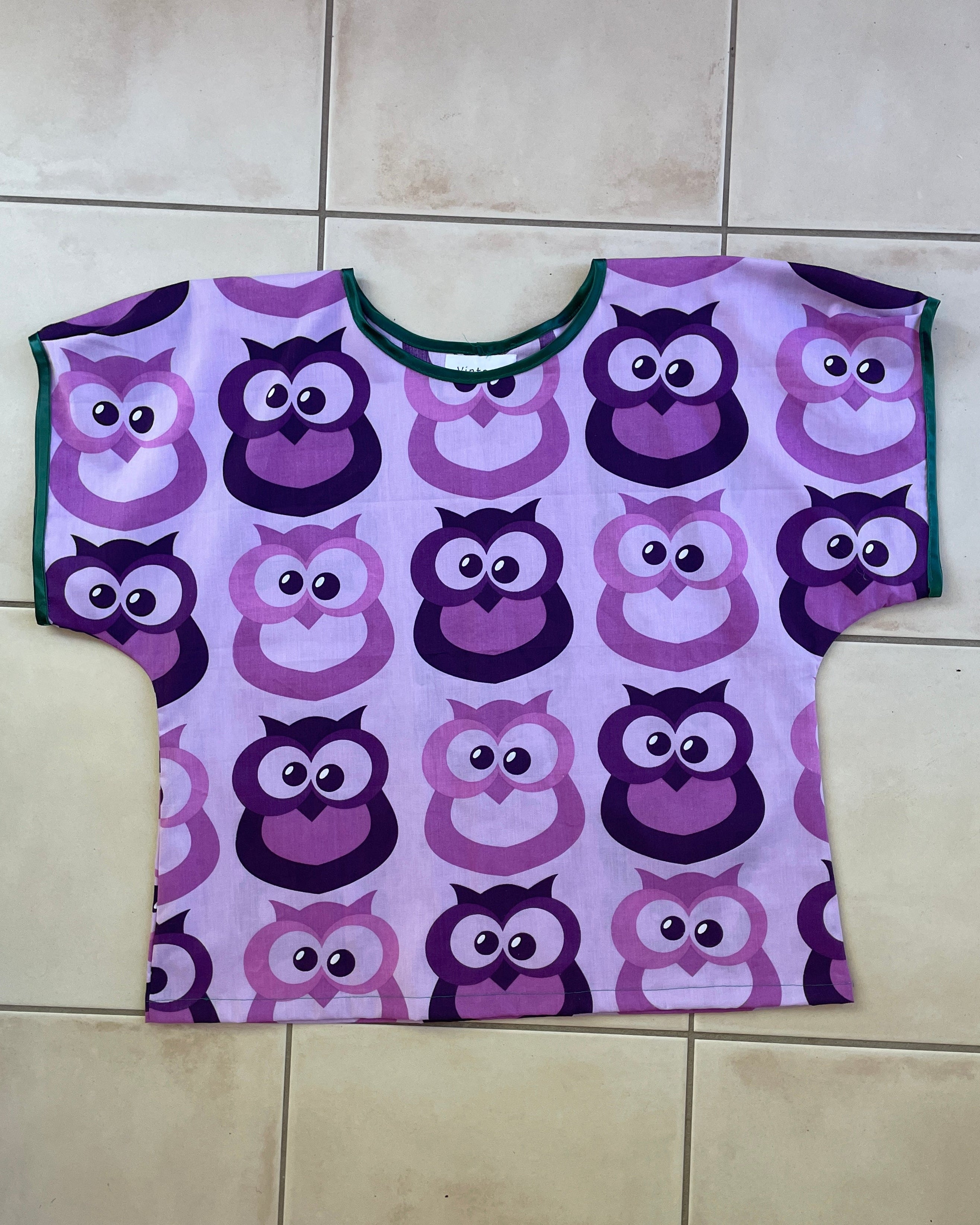 Purple t-shirt with owl pattern on a wooden hanger against a beige background