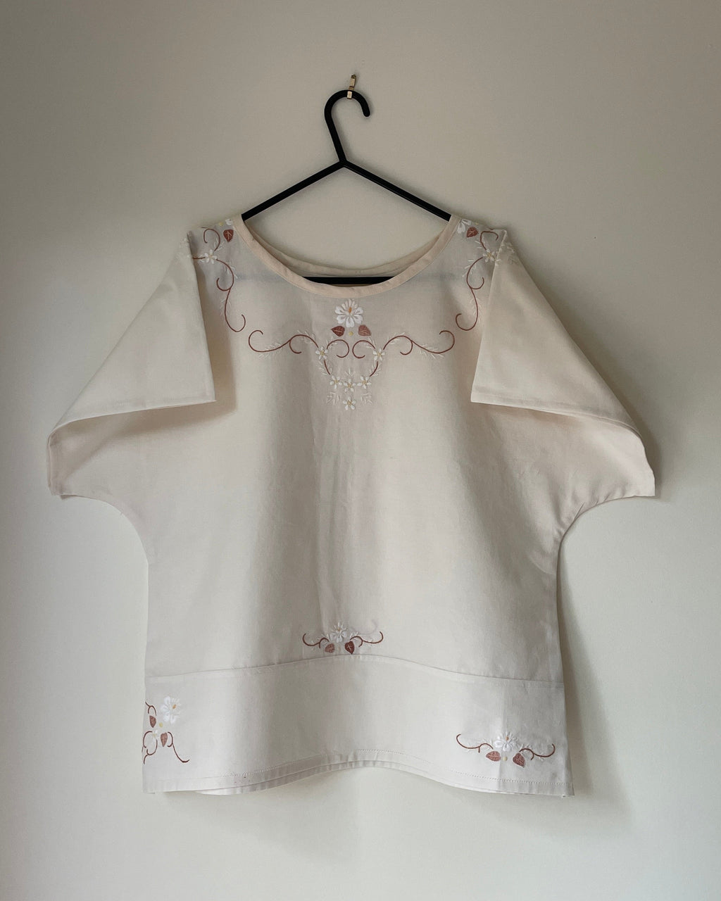White blouse with floral embroidery on a hanger against a plain wall.