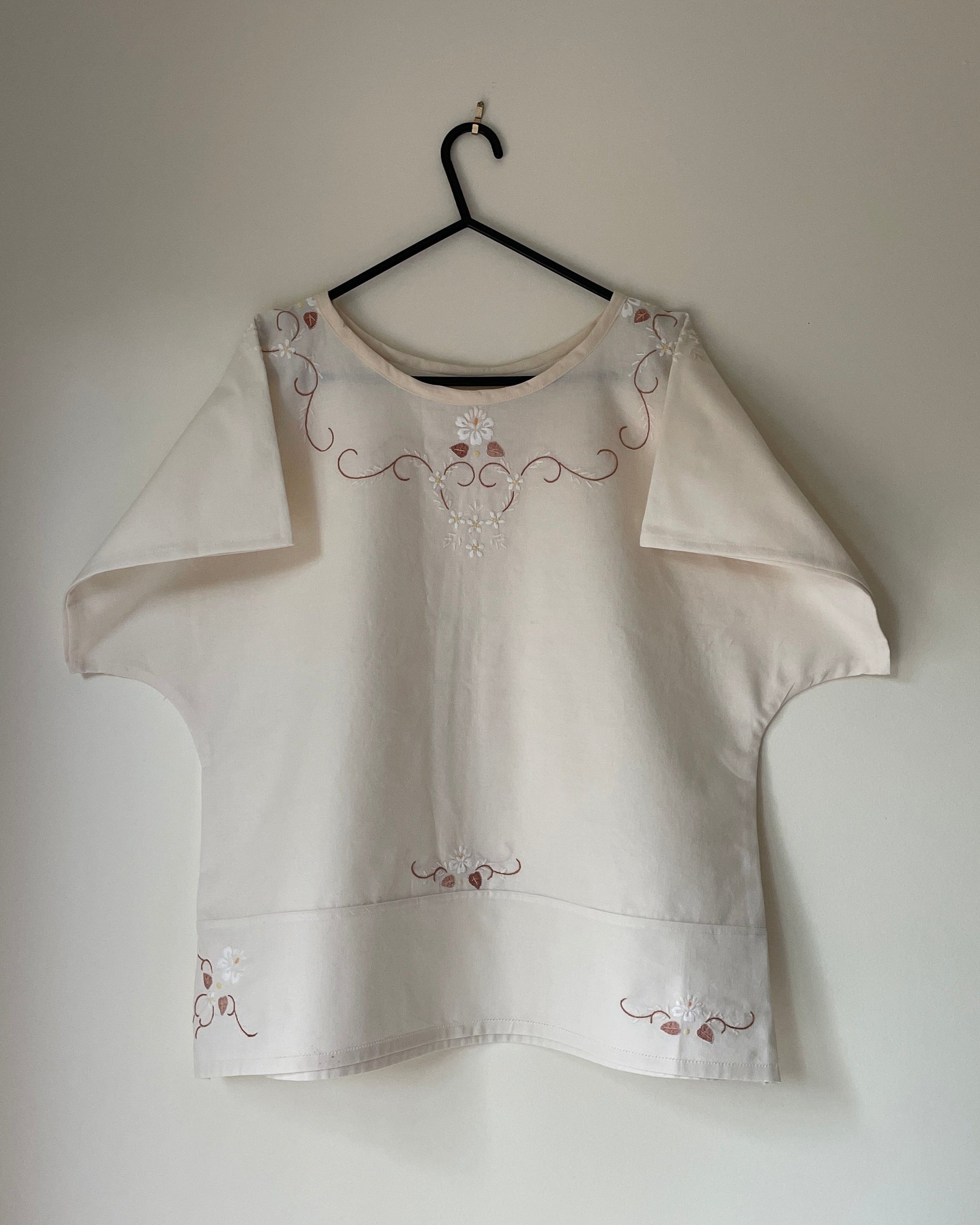 White blouse with floral embroidery on a hanger against a plain wall.