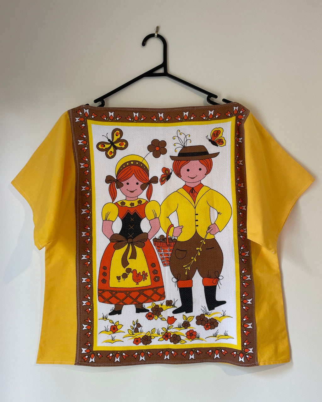 Yellow blouse with a printed design of two people and decorative elements on a hanger against a white wall.