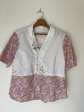 White blouse with floral sleeves and bottom on a wooden hanger against a white wall