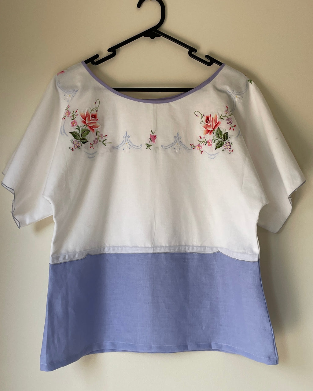 White blouse with floral embroidery and blue bottom on a hanger against a beige wall.