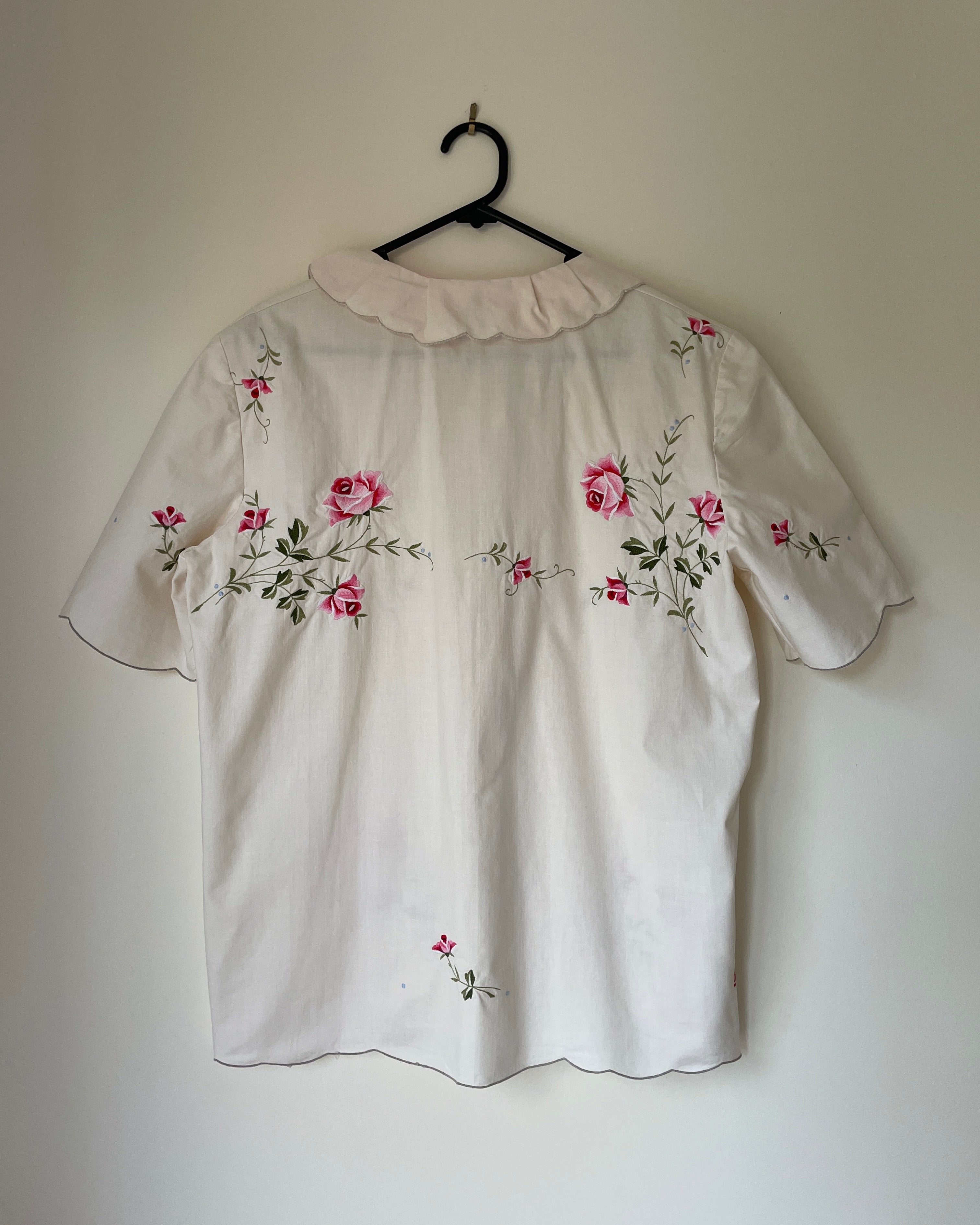 White blouse with floral embroidery on a hanger against a plain wall