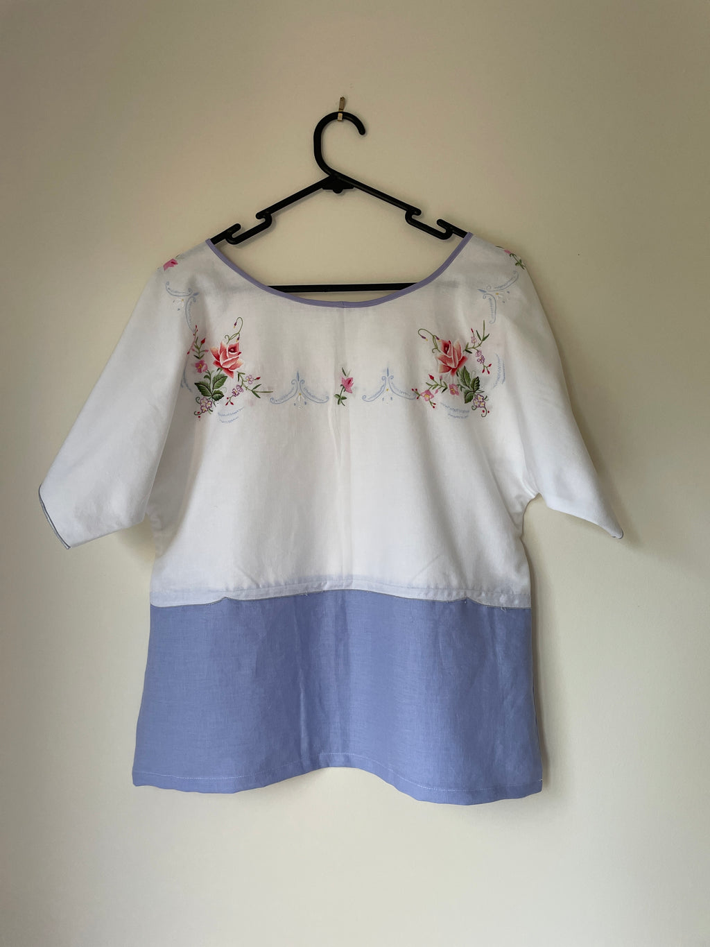 White blouse with floral embroidery and a blue hem on a hanger against a beige wall.