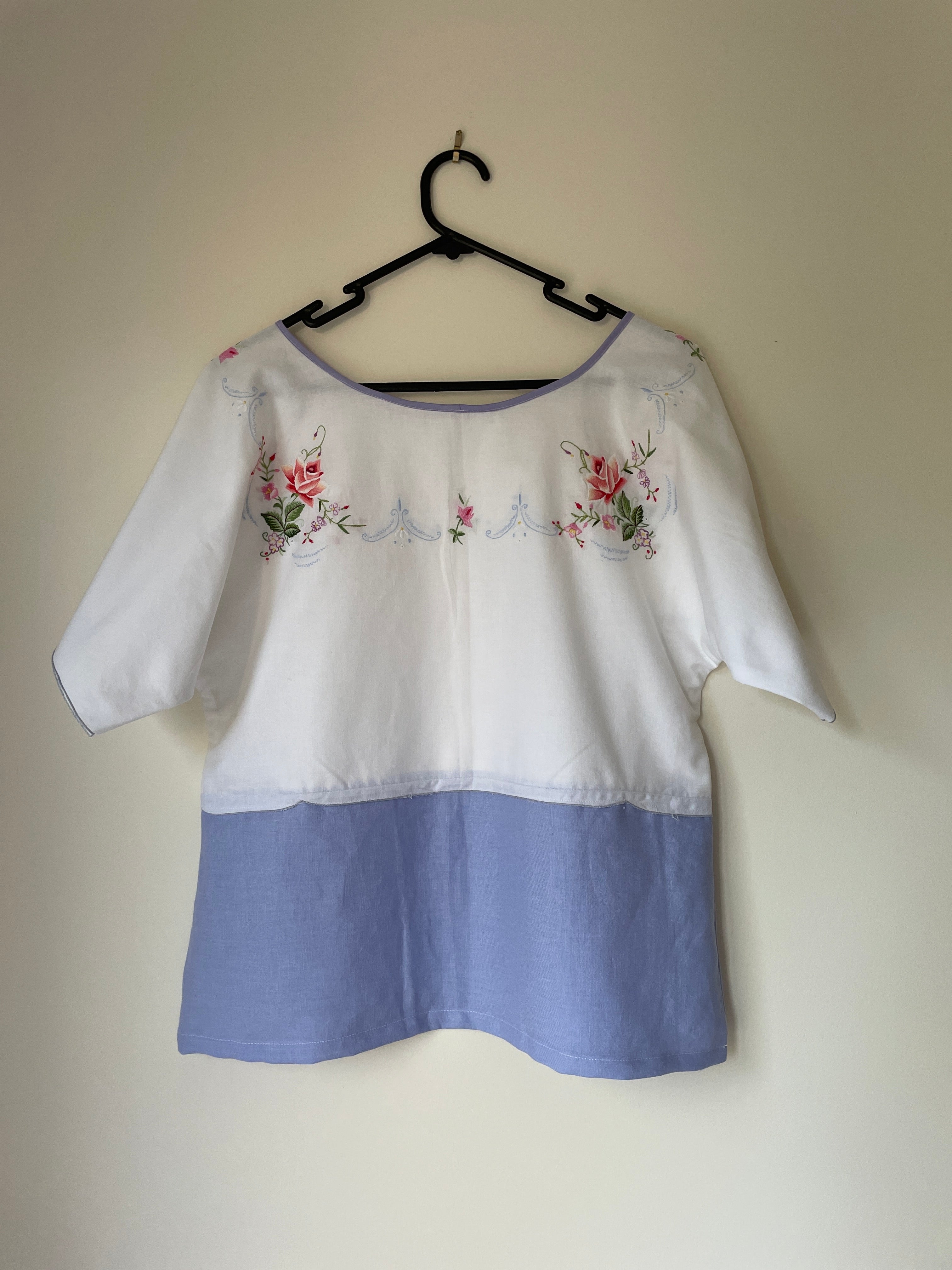 White blouse with floral embroidery and a blue hem on a hanger against a beige wall.