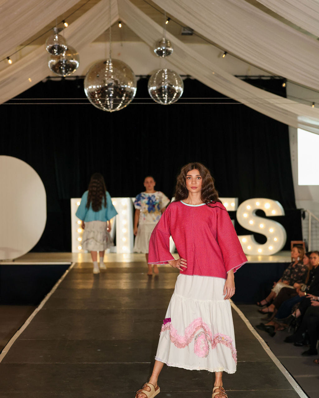 Model on a fashion show runway wearing a pink top and white skirt with floral details.