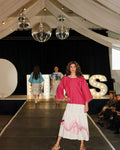 Model on a fashion show runway wearing a pink top and white skirt with floral details.