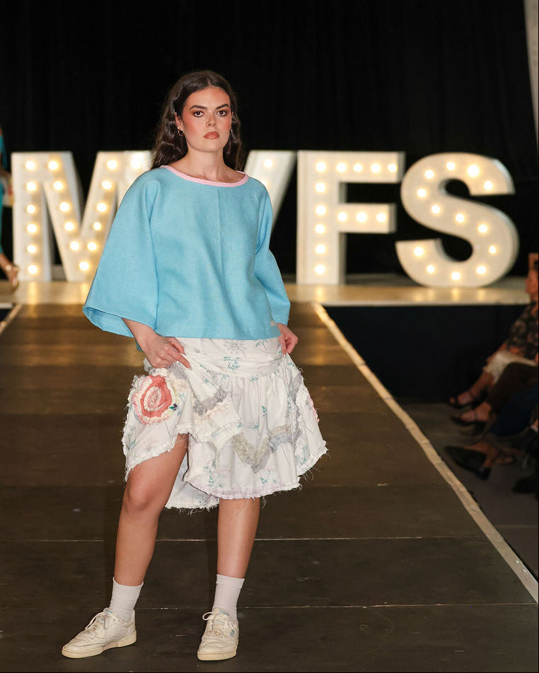 Model on a fashion show runway wearing a light blue top and floral skirt, with 'MVFS' branding in the background.