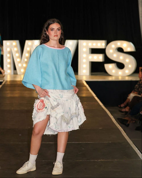Model on a fashion show runway wearing a light blue top and floral skirt, with 'MVFS' branding in the background.