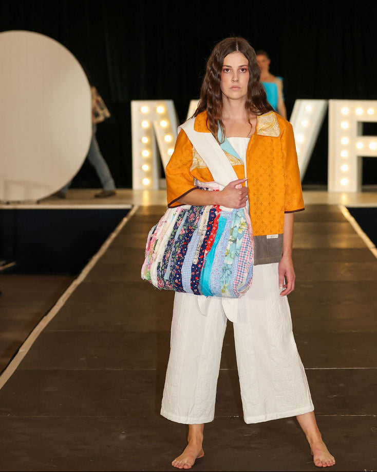 Model on a runway wearing an orange jacket and white pants, holding a colorful bag.