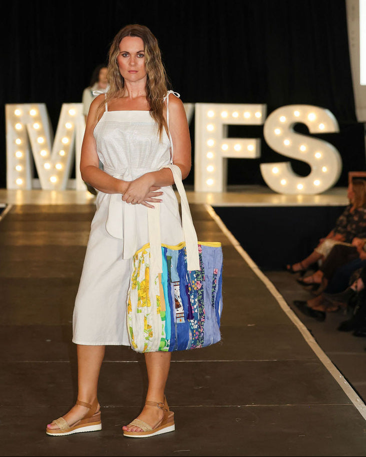 Model in a white dress holding a colorful bag on a runway with 'MVFS' letters in the background.
