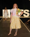 Model walking on a runway wearing a yellow dress with a pattern, with 'MVFS' illuminated letters in the background.