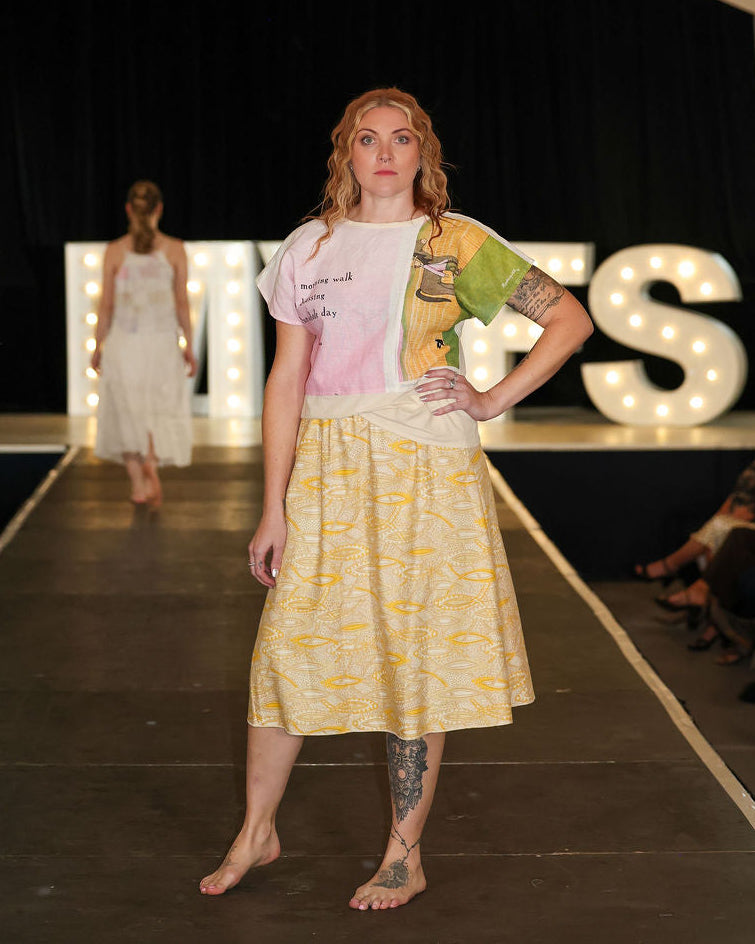 Model walking on a runway wearing a yellow dress with a pattern, with 'MVFS' illuminated letters in the background.