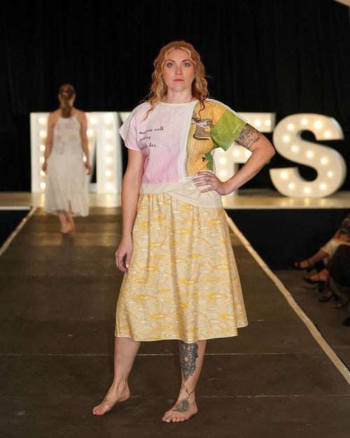 Model walking on a runway wearing a yellow dress with a pattern, with 'MVFS' illuminated letters in the background.