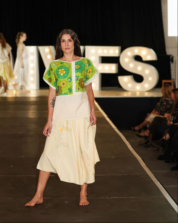 Model on a fashion runway wearing a green and white outfit with 'MVFS' branding in the background.