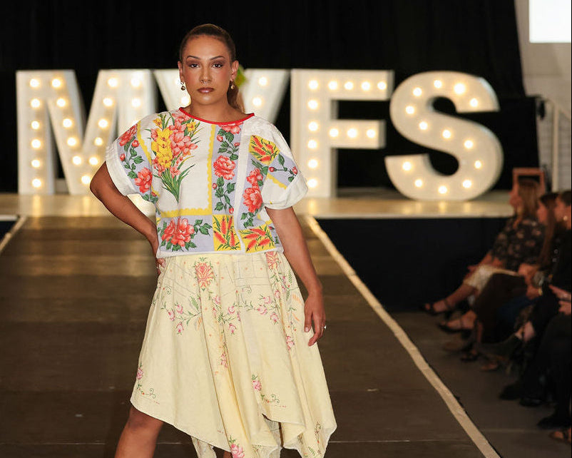 Model on a fashion runway wearing a colorful top and light skirt with 'MVFS' branding in the background.