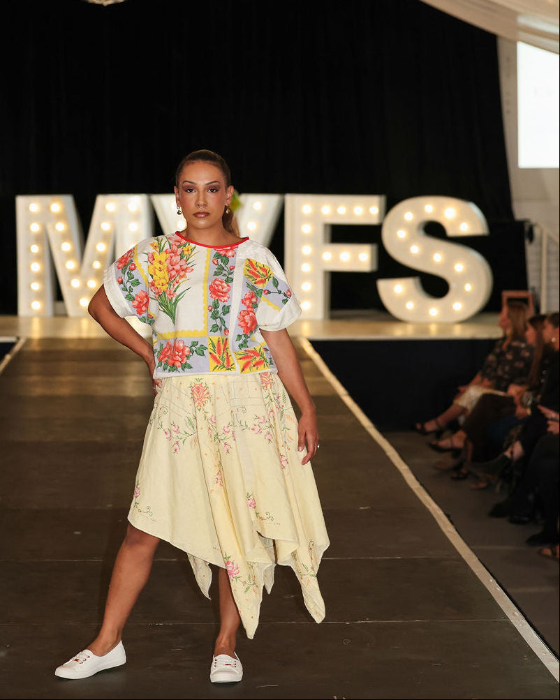 Model on a fashion runway wearing a colorful top and light skirt with 'MVFS' branding in the background.