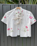 White embroidered blouse with pink flowers on a wooden hanger against a gray wooden fence.