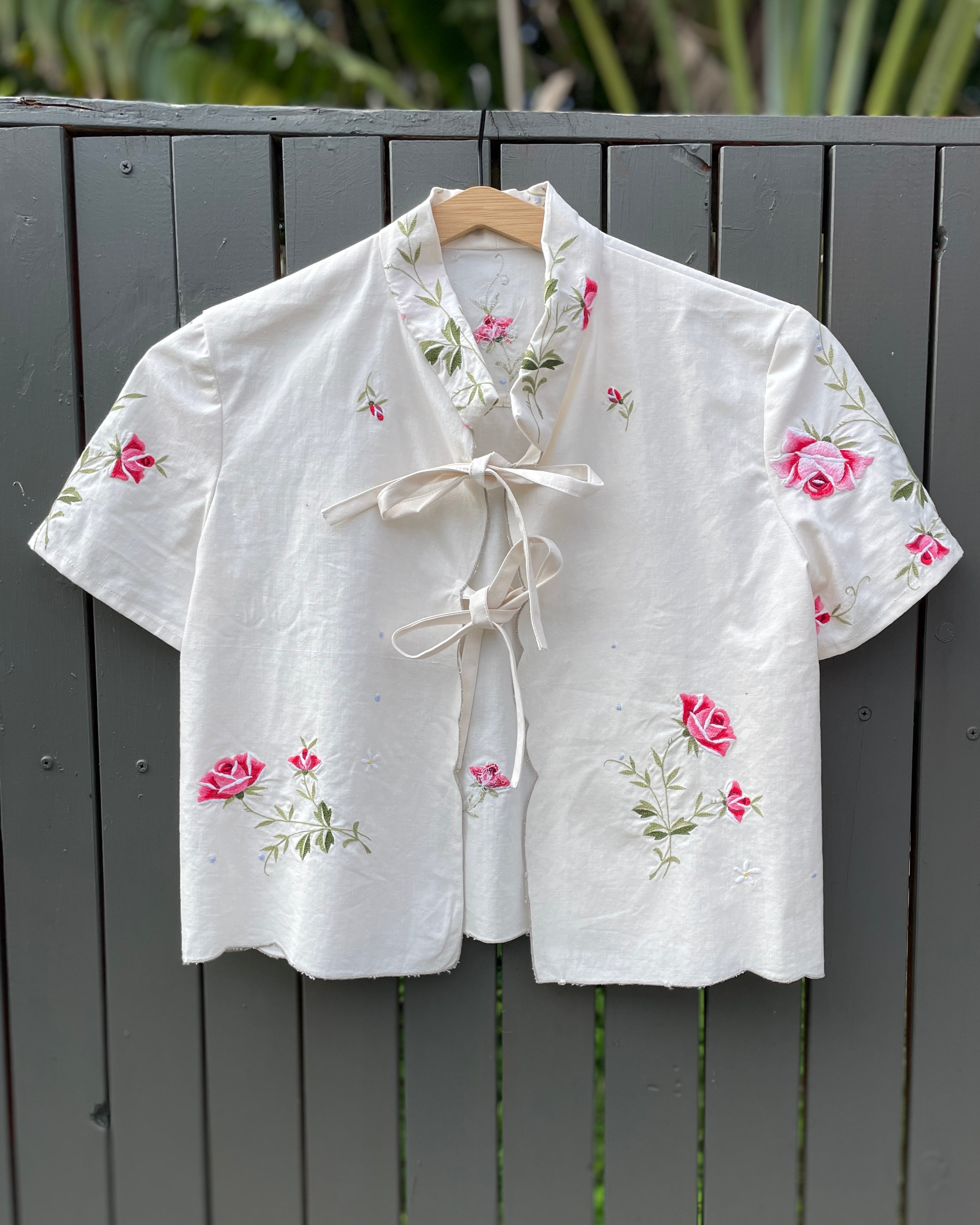 White embroidered blouse with pink flowers on a wooden hanger against a gray wooden fence.