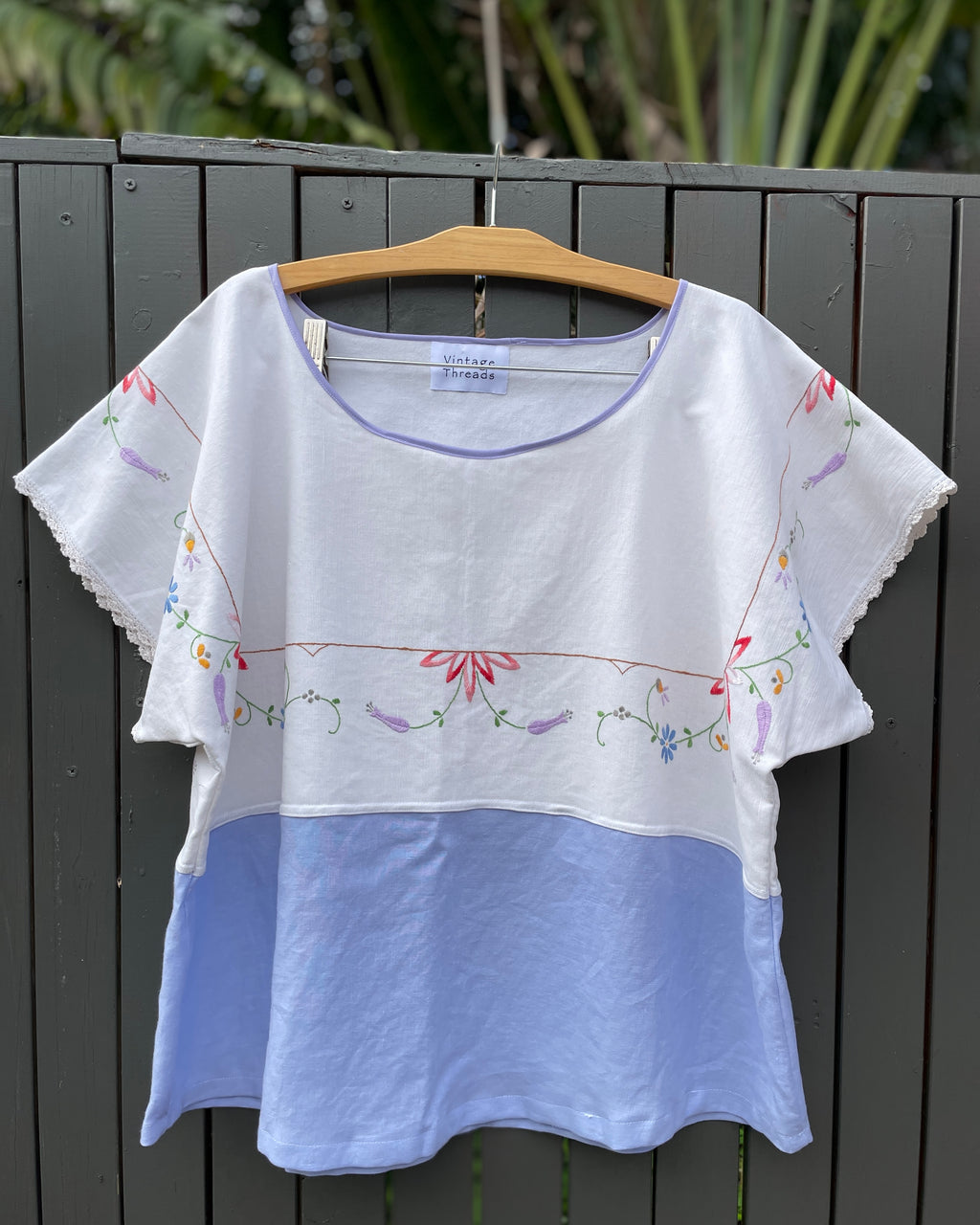 White blouse with floral embroidery on a hanger against a dark wooden fence.