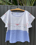 White blouse with floral embroidery on a hanger against a dark wooden fence.