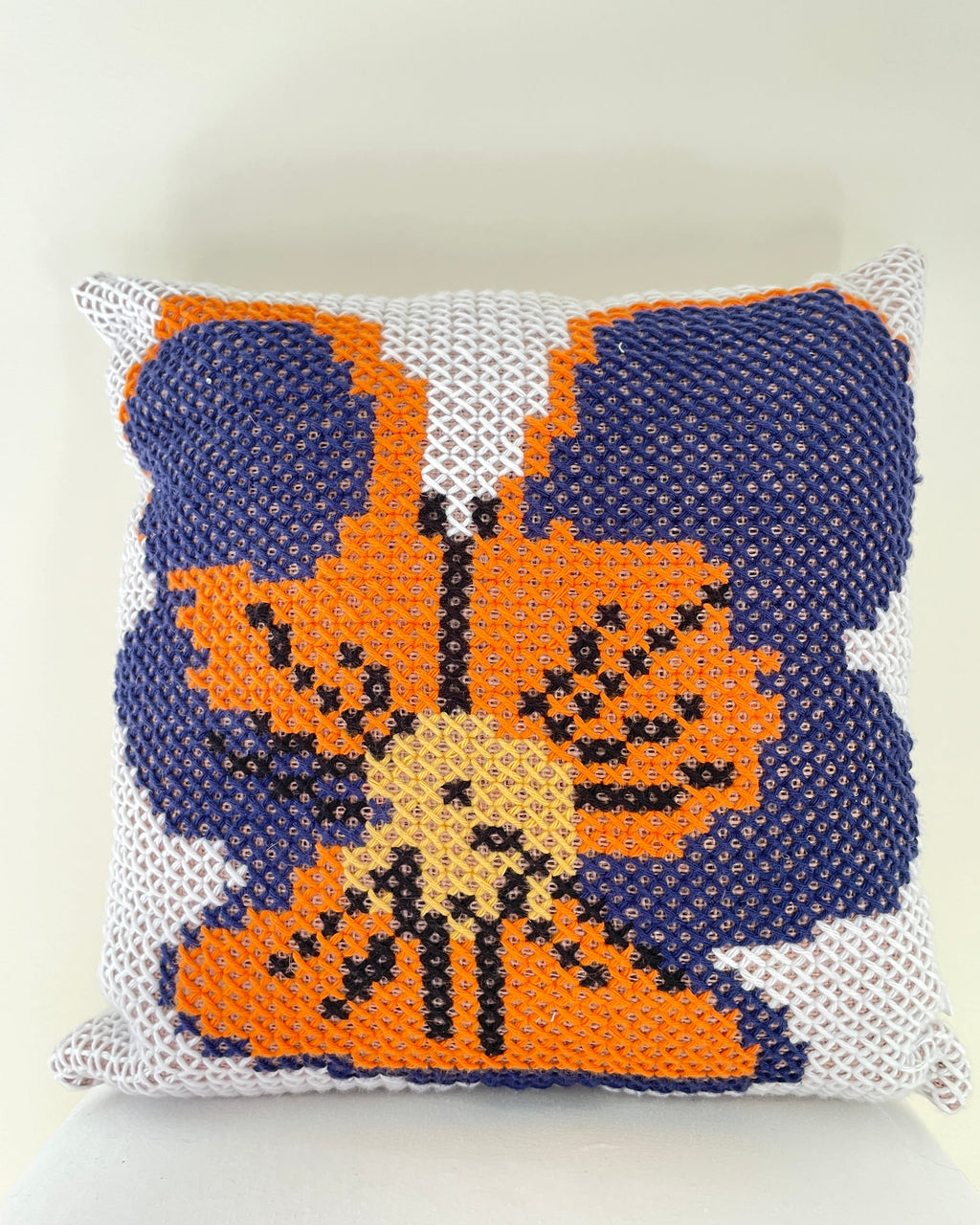 A square crocheted throw pillow featuring a pansy design in orange, navy blue, beige, and black on a white textured background.