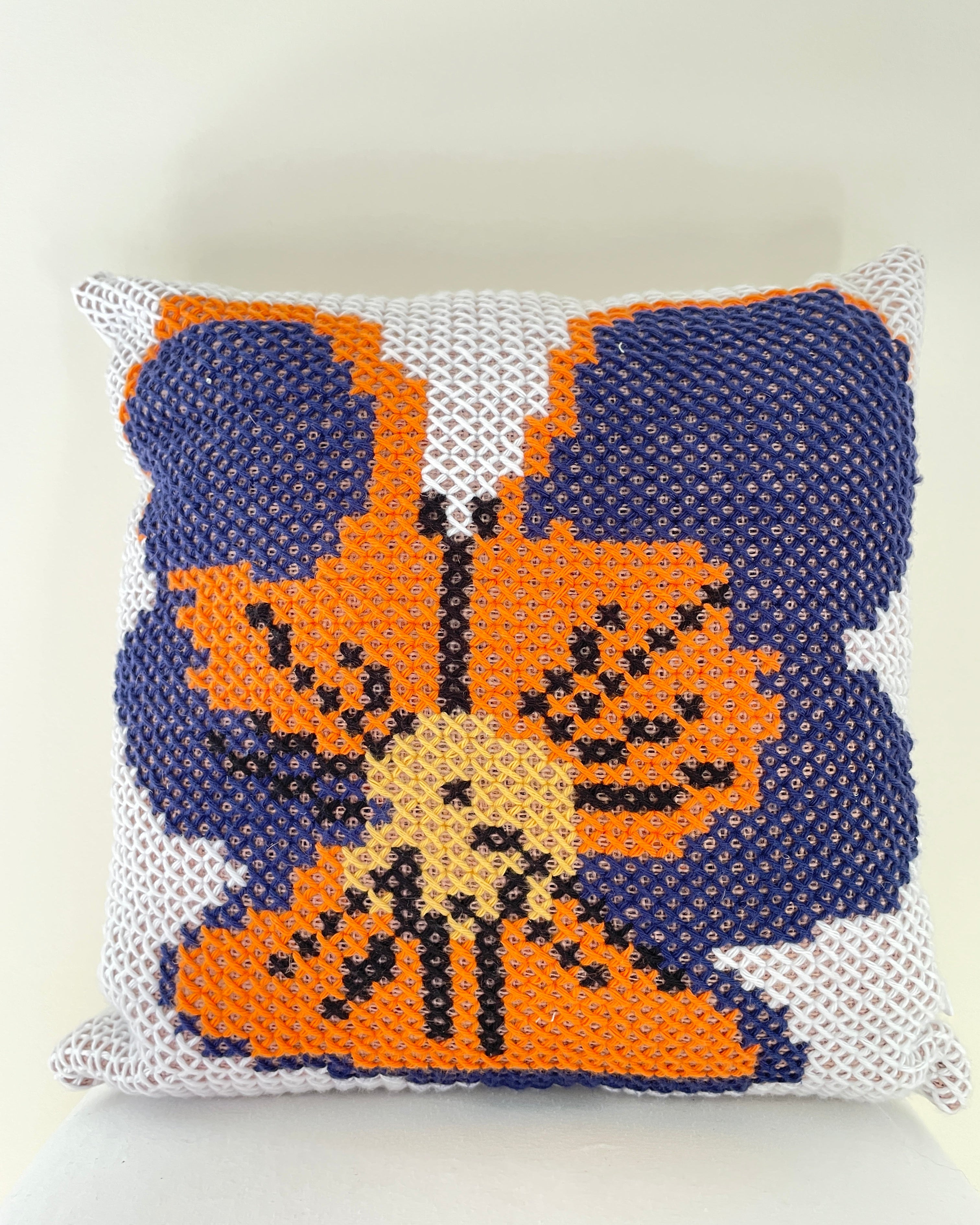 A square crocheted throw pillow featuring a pansy design in orange, navy blue, beige, and black on a white textured background.