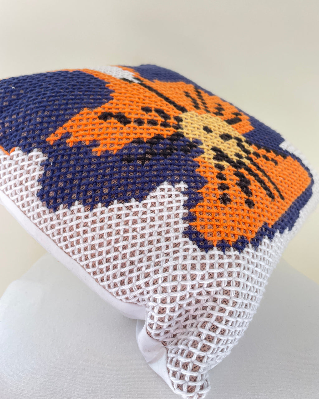 A close-up of a needlepoint cushion cover featuring a stylized pansy design in orange, navy blue, and beige, with a white mesh backing.
