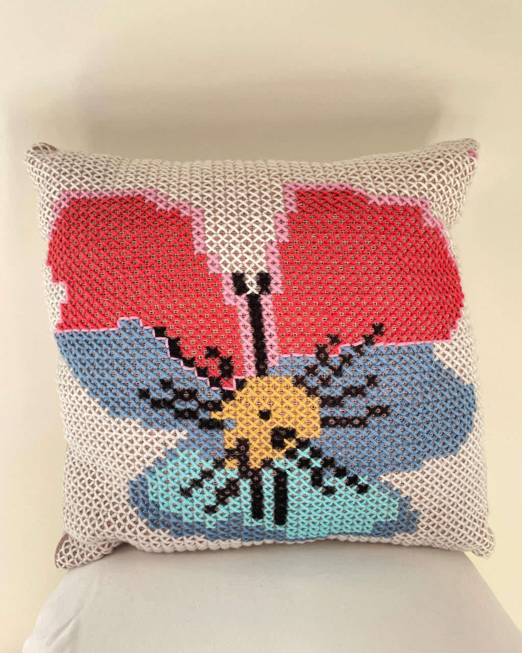 A square cross stitch throw pillow featuring a stylized flower design with red, light blue, and yellow sections on a beige knit background, resting on a white surface against a light beige wall.