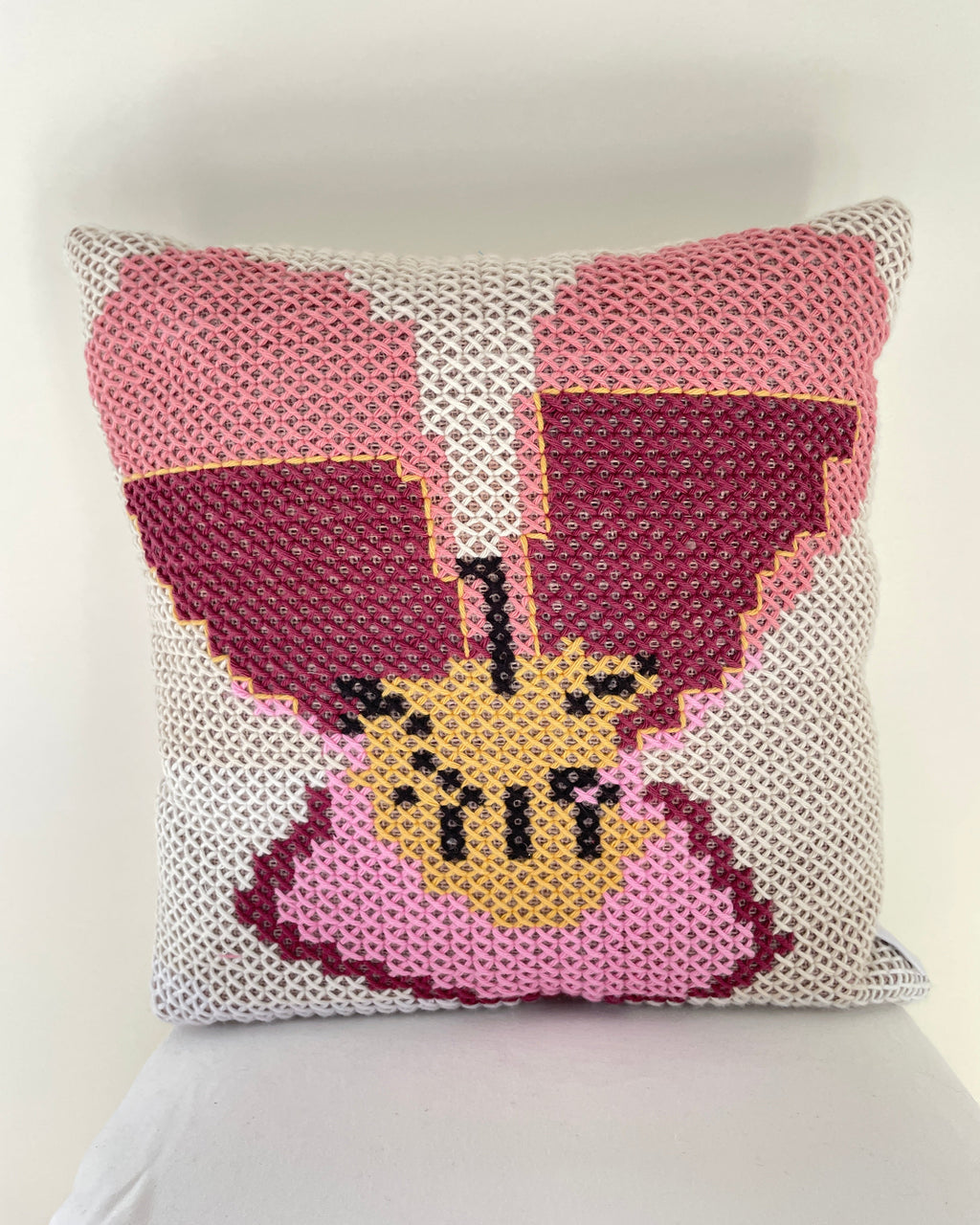 A square cross stitch throw pillow featuring a stylized pansy design in shades of pink, burgundy, beige, and black on a white knit background.