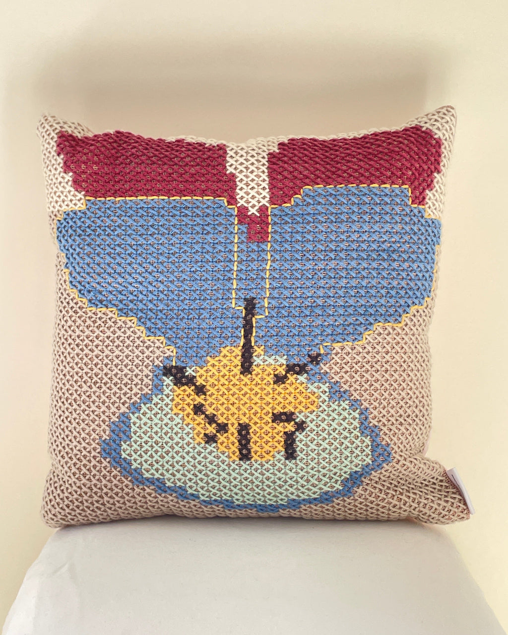 A square cross stitch throw pillow with a stylized floral design in blue, burgundy, yellow, and light blue on a beige textured background, resting against a white pillow on a light beige surface.