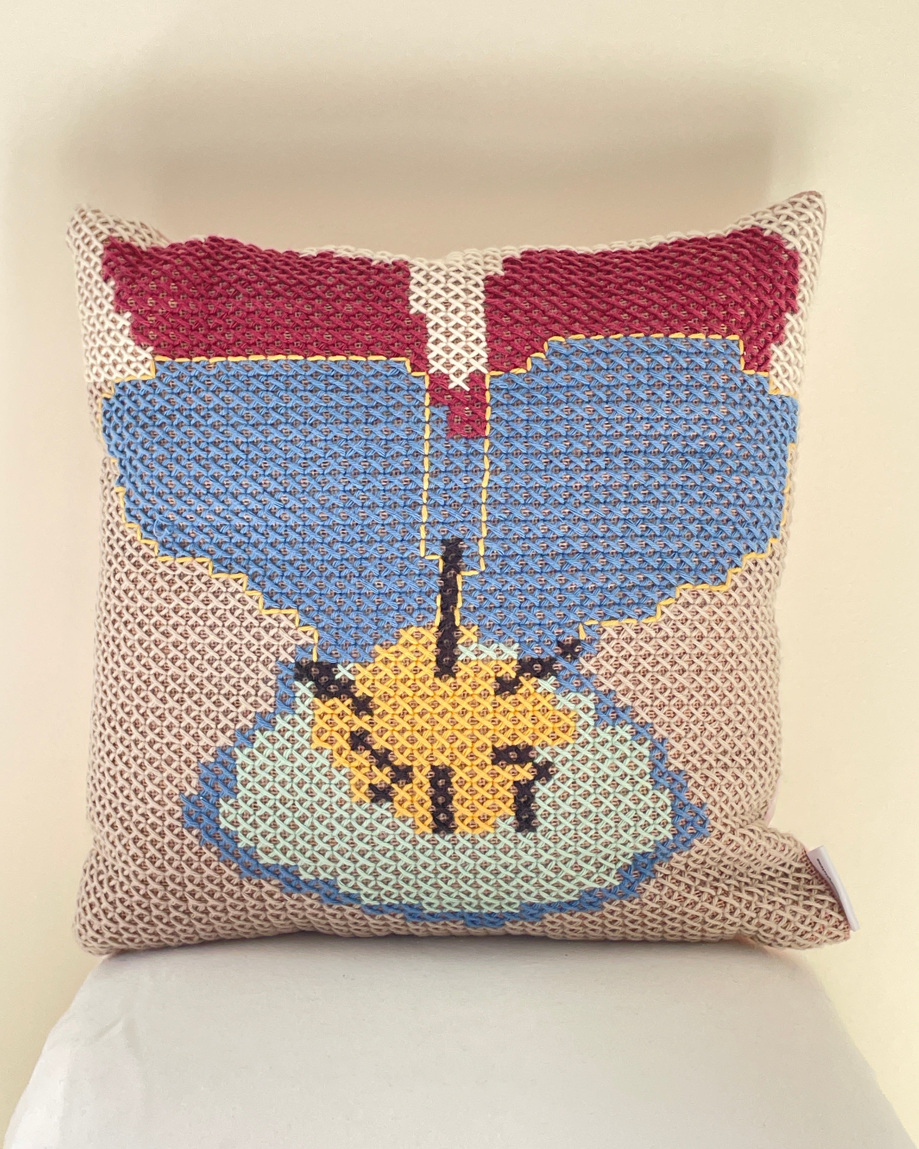 A square cross stitch throw pillow with a stylized floral design in blue, burgundy, yellow, and light blue on a beige textured background, resting against a white pillow on a light beige surface.