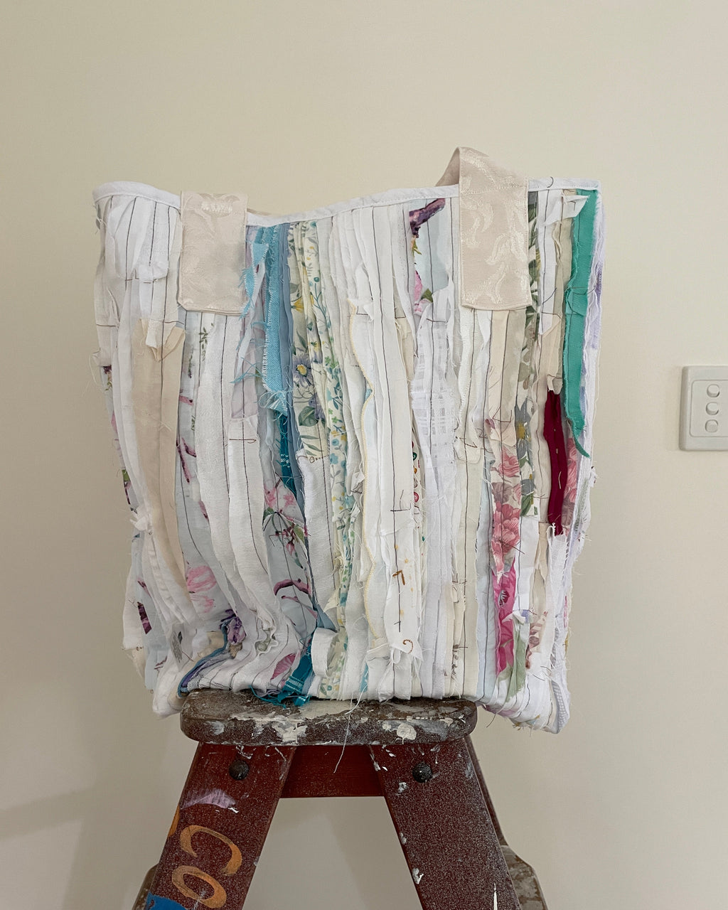 A handmade tote bag made from vertical strips of mixed vintage fabrics in white, beige, blue, pink, and green, displayed on a wooden ladder against a plain beige wall.