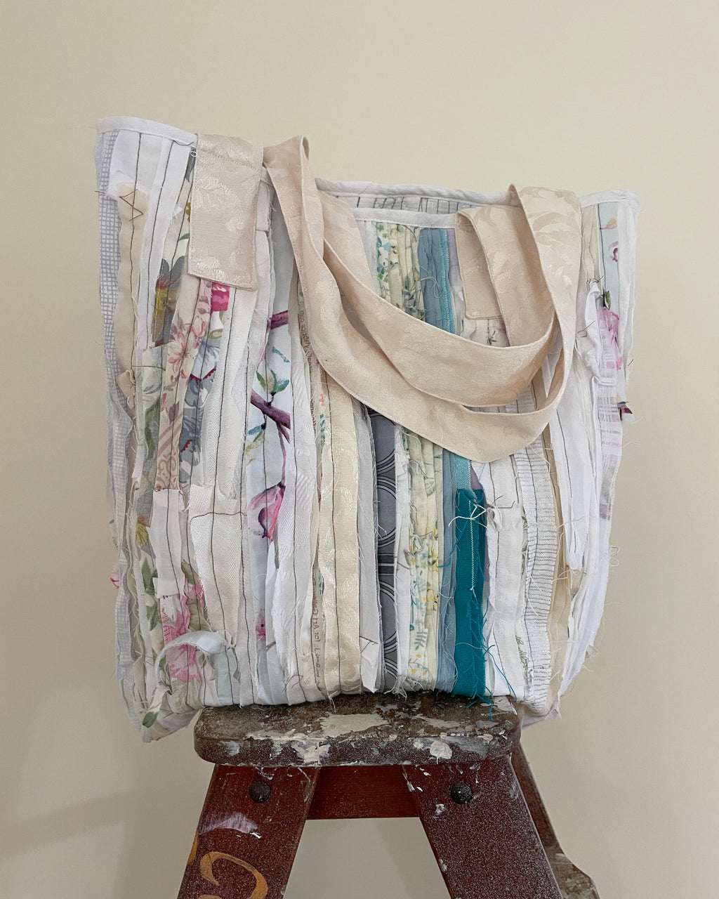 A handmade tote bag made from vertical strips of vintage floral and patterned fabric, featuring beige canvas shoulder straps, displayed on a worn wooden stool against a plain beige wall.