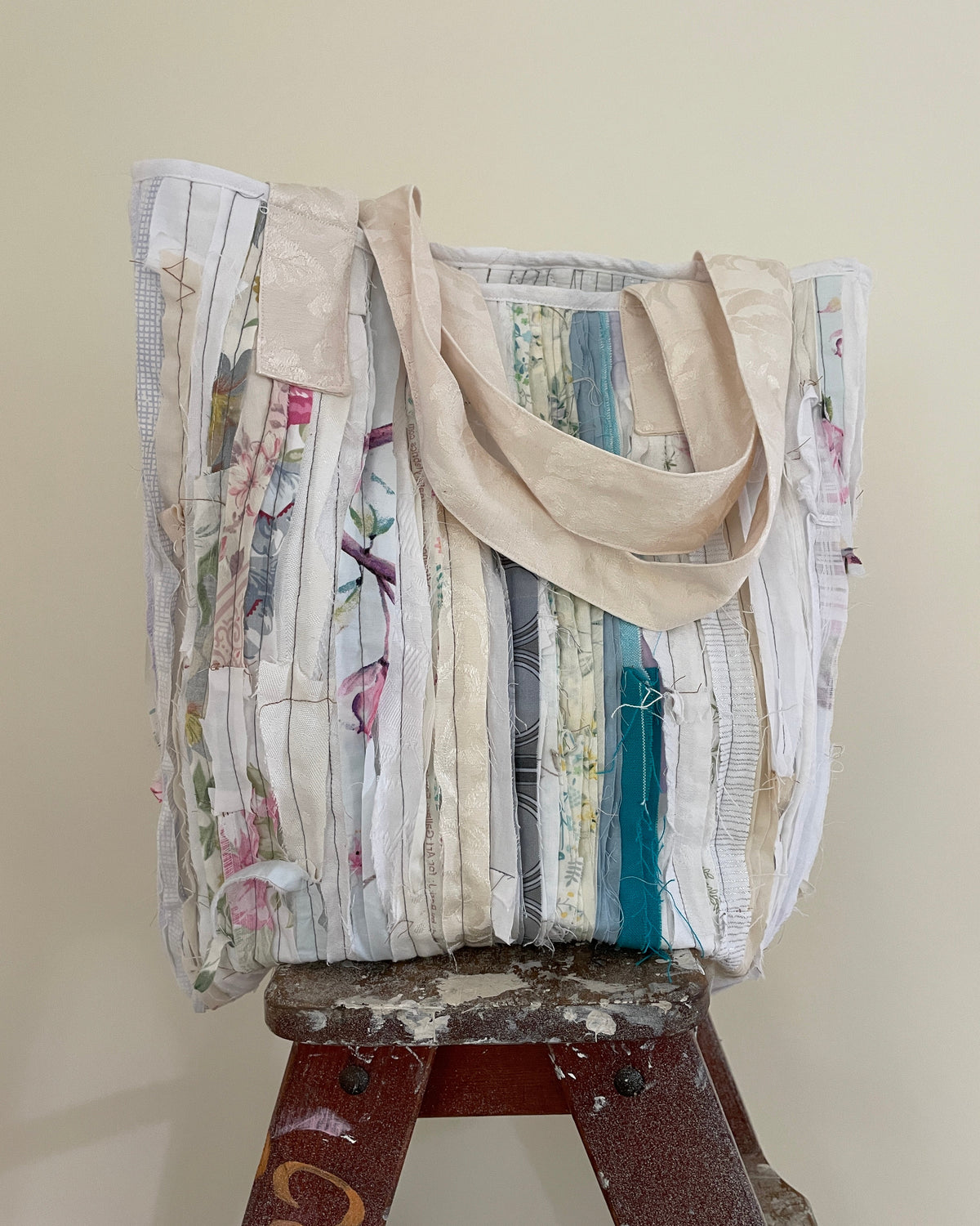 A handmade tote bag made from vertical strips of vintage floral and patterned fabric, featuring beige canvas shoulder straps, displayed on a worn wooden stool against a plain beige wall.