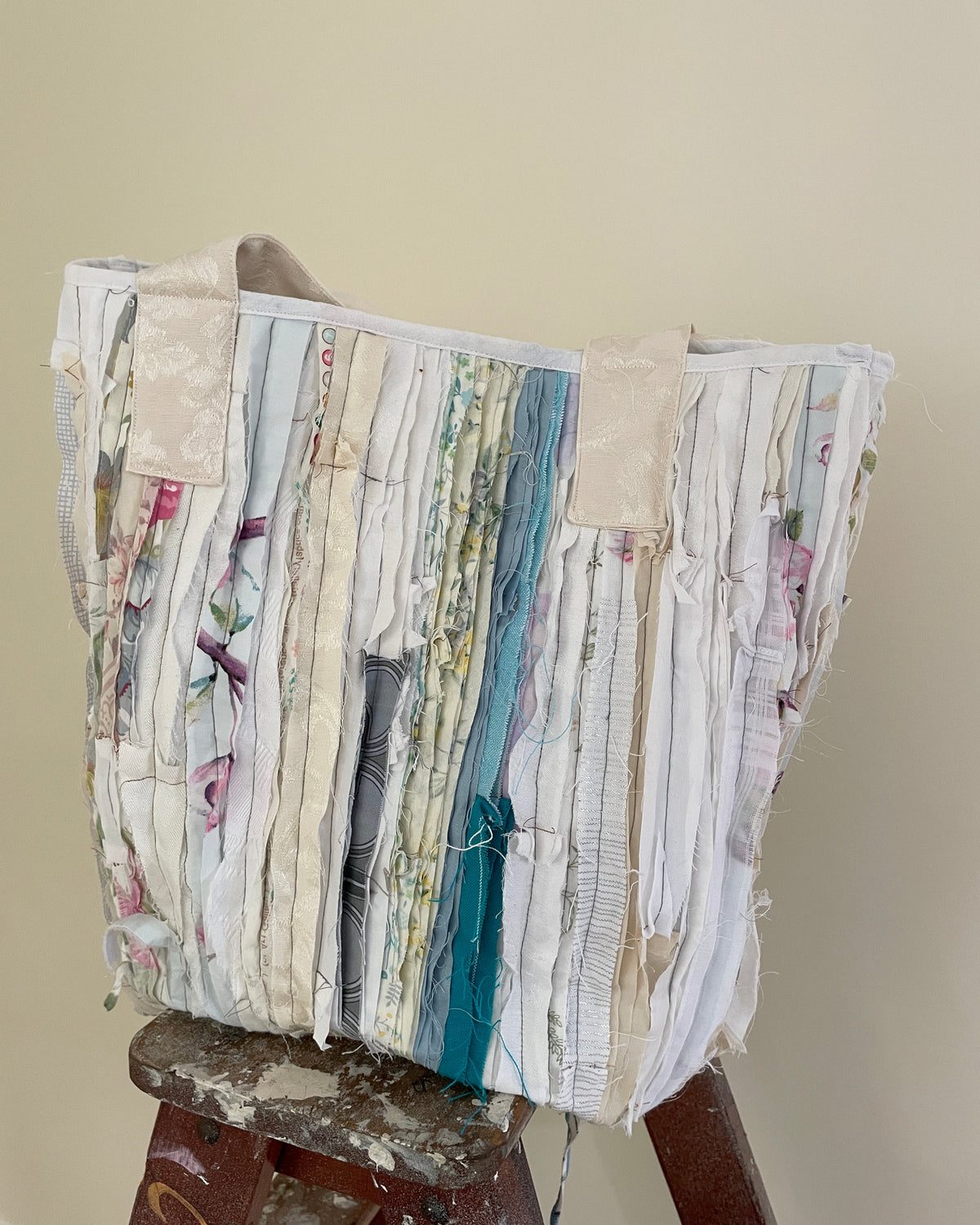 A handmade tote bag made from vertical strips of mixed fabric, including floral prints, silks, and solid colors, displayed on a wooden stool against a plain beige wall.