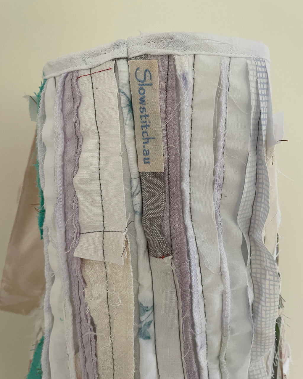 A close-up of a handmade fabric bag or pouch made from vertical strips of mixed textiles in soft neutral and pastel tones, including cream, beige, light purple, and gray. A fabric label reading 'Slow stitch au' is stitched onto the front.