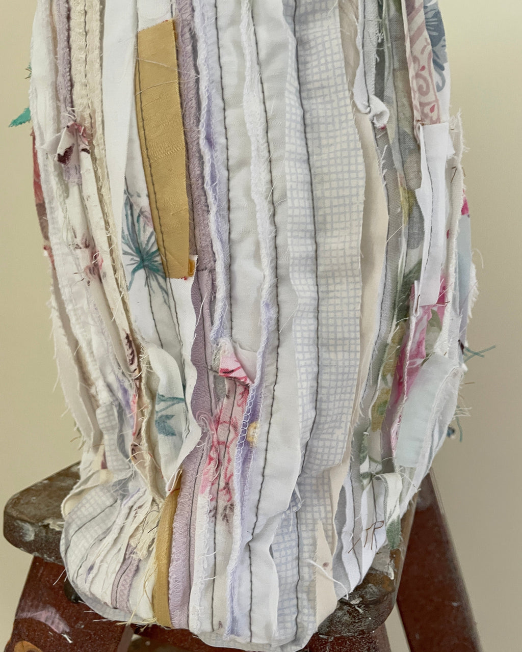 A close-up of a handmade fabric bag or pouch made from vertical strips of mixed vintage and thrifted textiles in soft pastel colors, including cream, lavender, pink, and light blue, with a small tan patch on the front, resting on a wooden stool.
