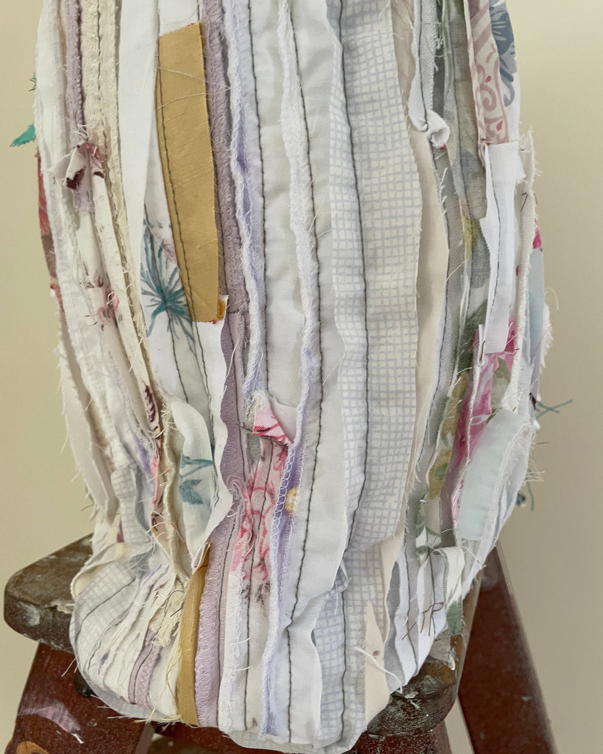 A close-up of a handmade fabric bag or pouch made from vertical strips of mixed vintage and thrifted textiles in soft pastel colors, including cream, lavender, pink, and light blue, with a small tan patch on the front, resting on a wooden stool.