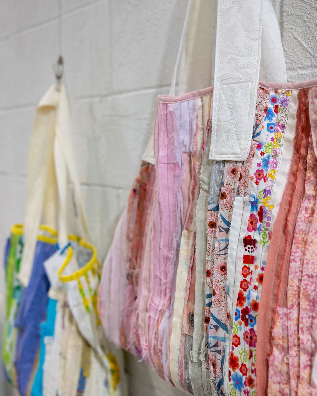 A close-up of several fabric tote bags hanging from hooks against a light gray brick wall. The bags feature mixed prints and textures, including floral patterns, pastel pink tones, and cream fabric panels.