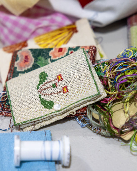 A close-up of a handmade embroidered fabric sampler with a floral pattern and a small cross-stitch design, surrounded by colorful threads, tags, and sewing materials on a light surface.