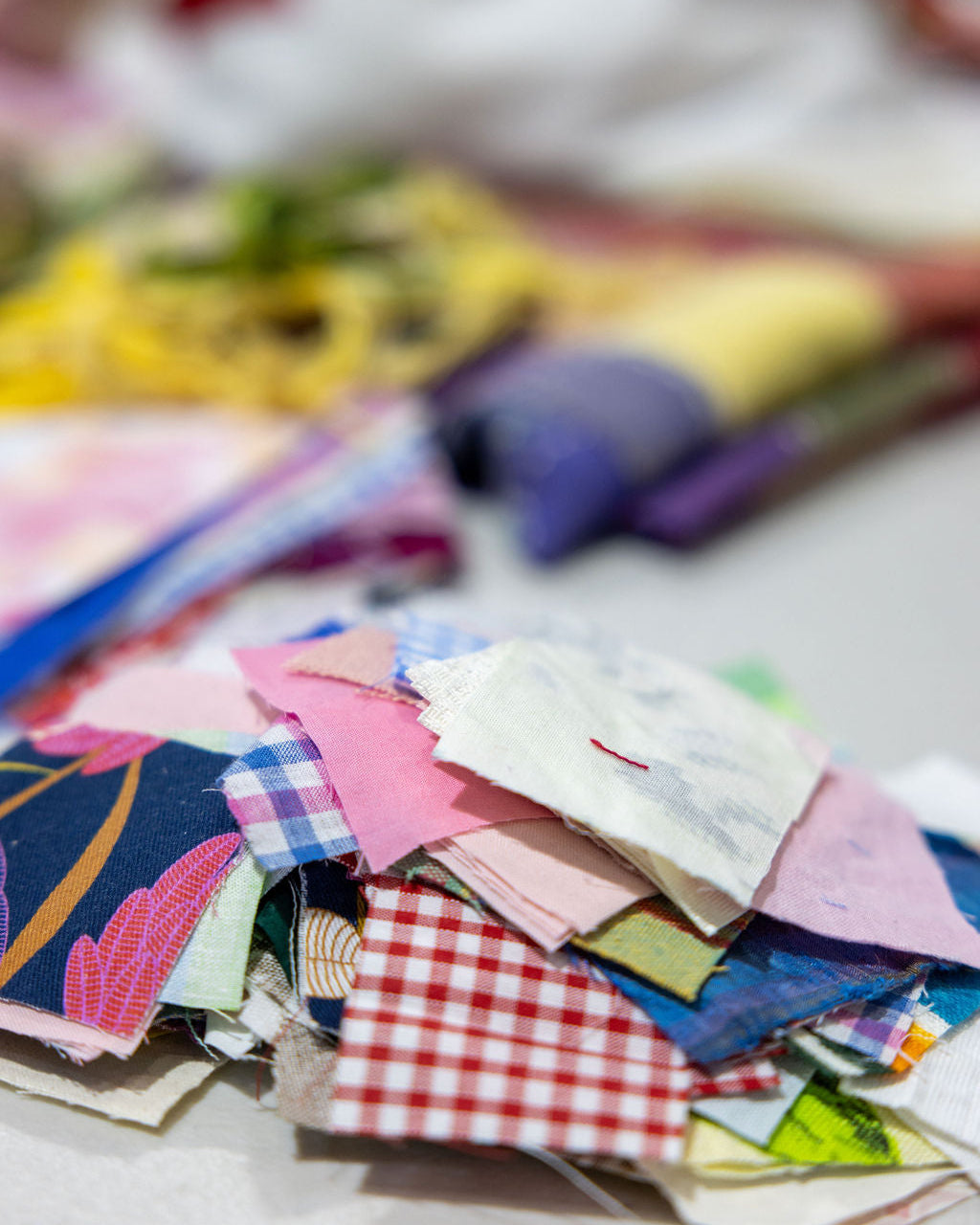 A pile of colorful fabric swatches in various patterns and colors, including gingham, plaid, floral, and solid prints, arranged on a light surface with a blurred background.