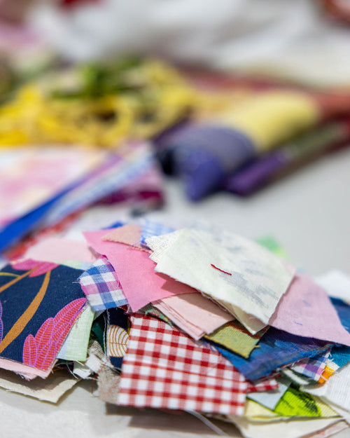 A pile of colorful fabric swatches in various patterns and colors, including gingham, plaid, floral, and solid prints, arranged on a light surface with a blurred background.