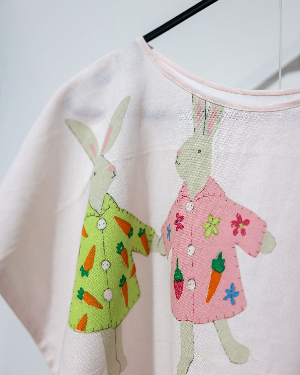 A close-up of a light pink short-sleeve top hanging on a black hanger, featuring two printed bunny characters wearing colorful dresses decorated with carrots and flowers.